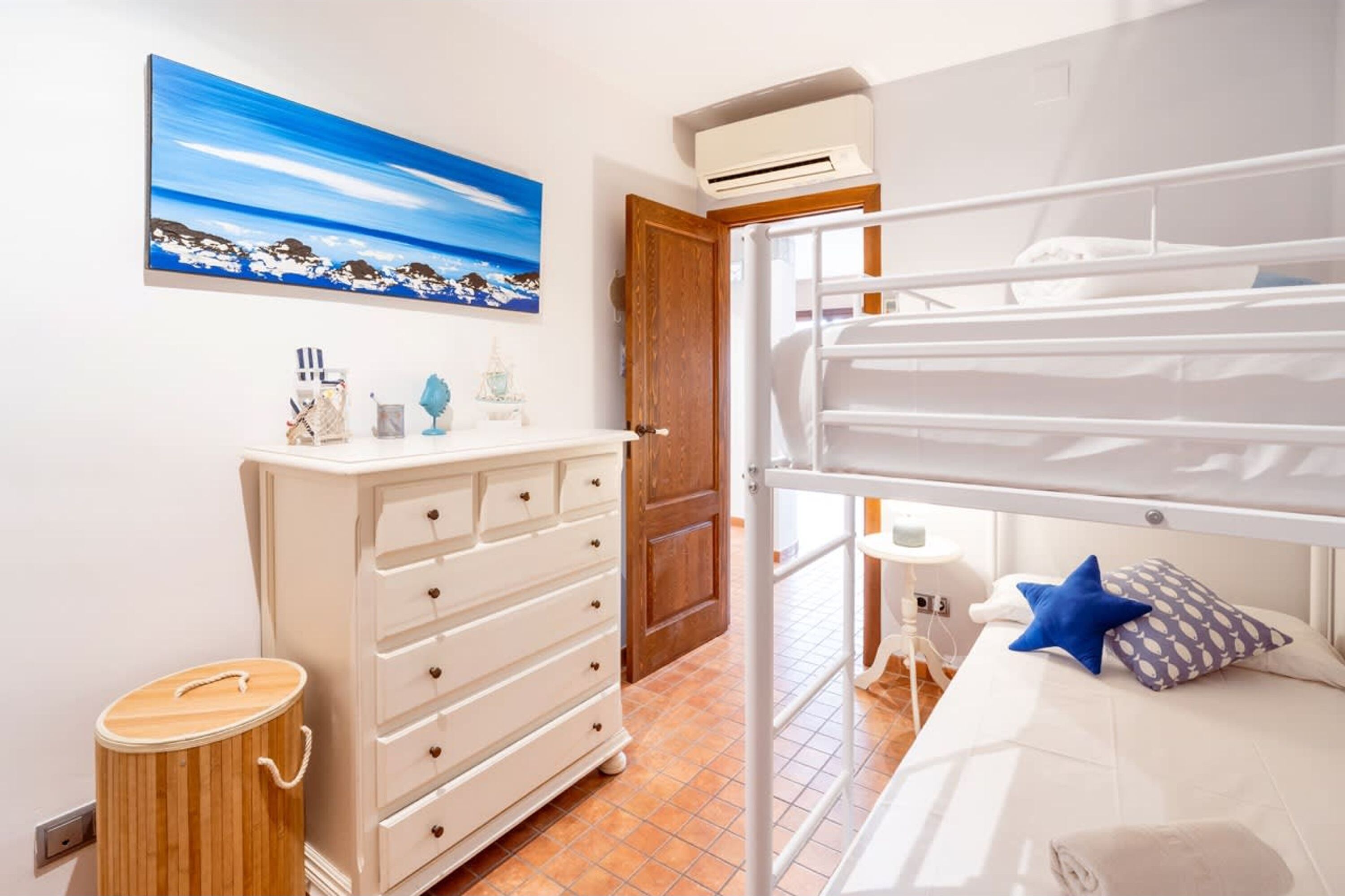 Quarto de hotel com cama beliche, penteadeira, um banquinho de madeira e uma pintura de paisagem costeira.