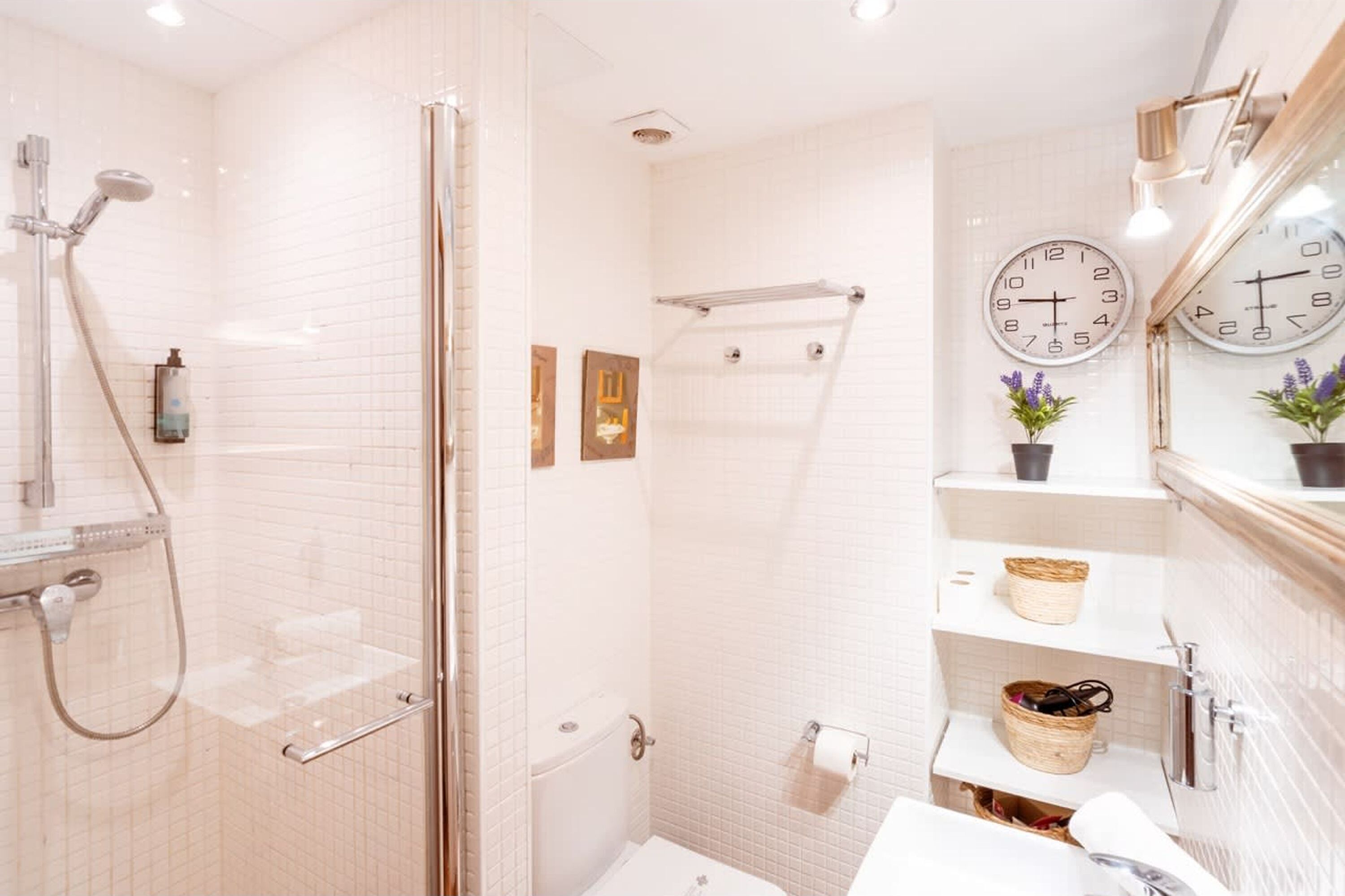 Un baño moderno con cabina de ducha de vidrio, paredes cubiertas de azulejos blancos, dos relojes y un pequeño estante con cestas.
