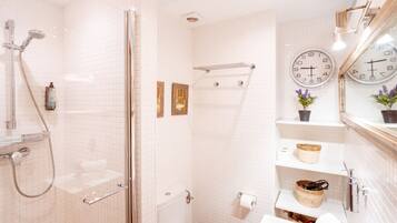 Un baño moderno con cabina de ducha de vidrio, paredes cubiertas de azulejos blancos, dos relojes y un pequeño estante con cestas.