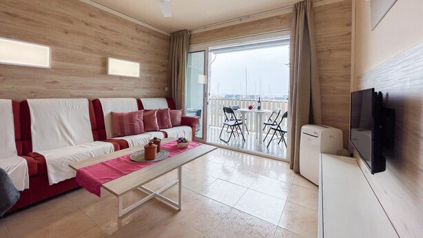 A modern living room with a sofa, a coffee table, and a TV. There is a balcony with chairs and a table. - Unik Vacation Marina (Tarragona)