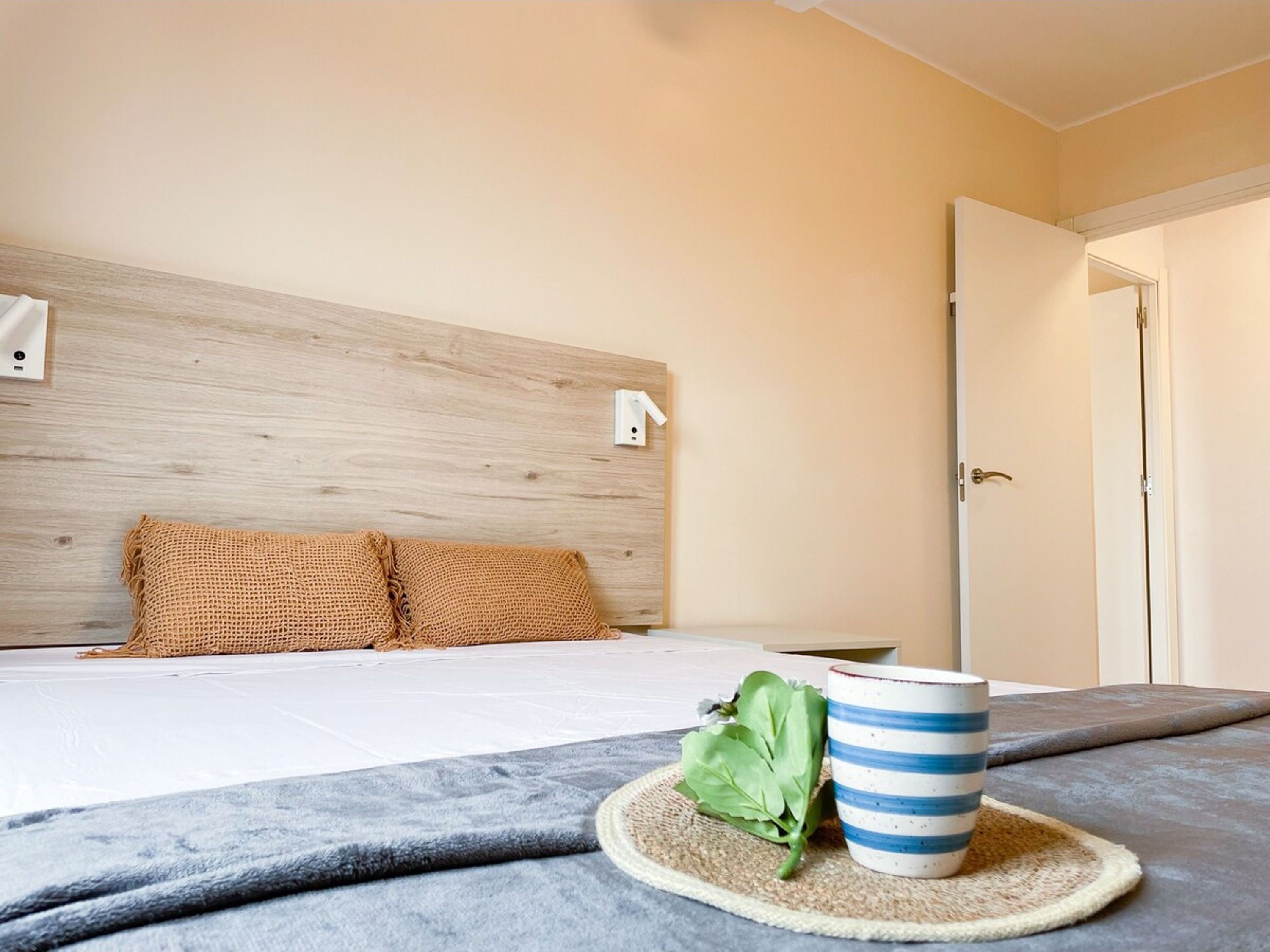 Una cama bien hecha con cabecera, una taza azul a rayas sobre un posavasos y una puerta a la derecha.