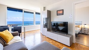 A modern living room with a large TV, a sofa, a dining table, and a view of the ocean. - Unik Vacation Via Augusta (Tarragona)