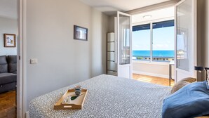 A bedroom with a bed, a bedside table with a tray, a framed picture, and a view of the sea through an open door. - Unik Vacation Via Augusta (Tarragona)