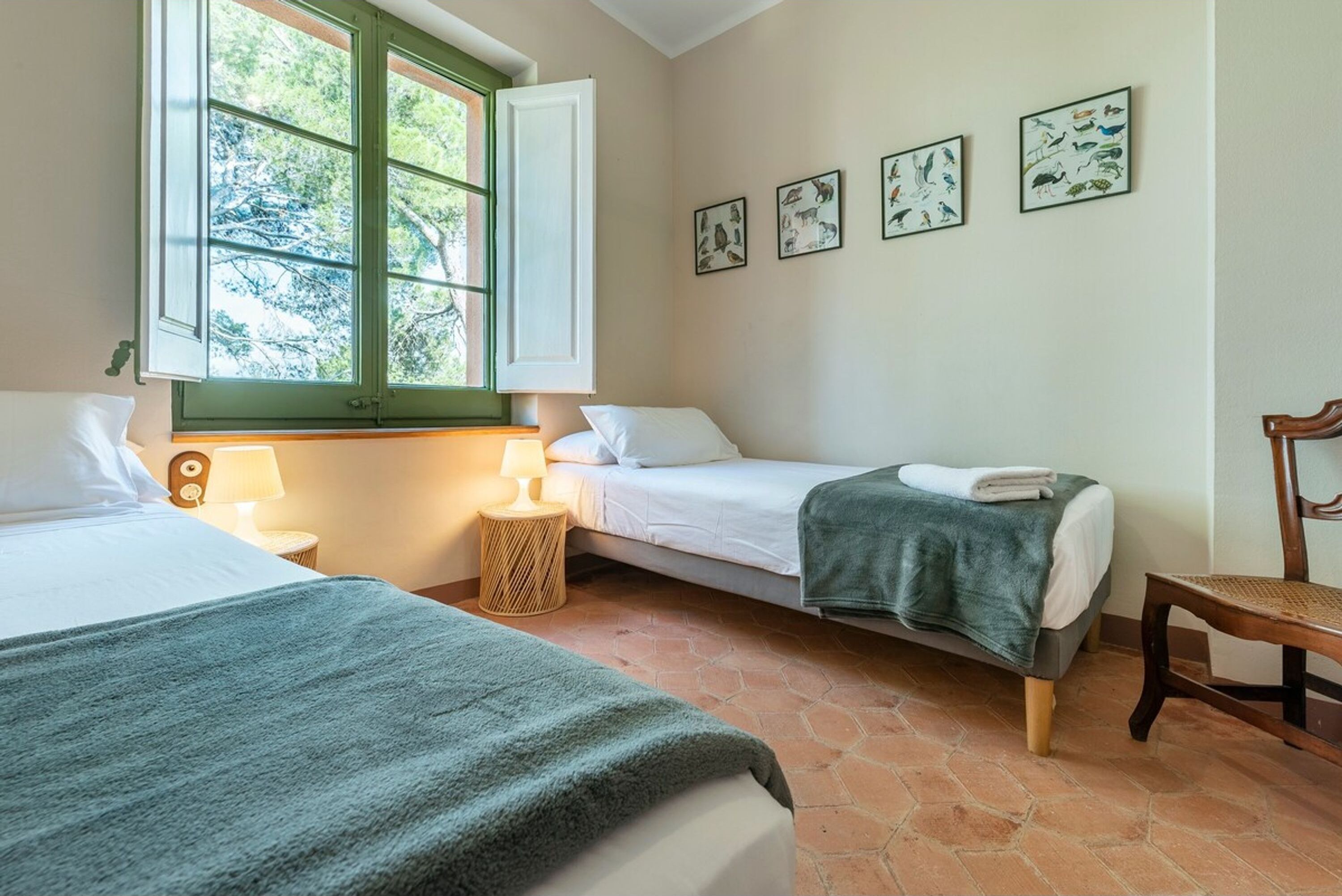 Habitació amb dos llits, una finestra amb persianes verdes, una làmpada i decoració mural amb representació d'animals.