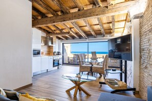 A modern living room with a glass coffee table, wicker furniture, and a TV. A kitchen with white cabinets and a dining area with wicker chairs are visible. - Unik Vacation Panoramic (Tarragona)