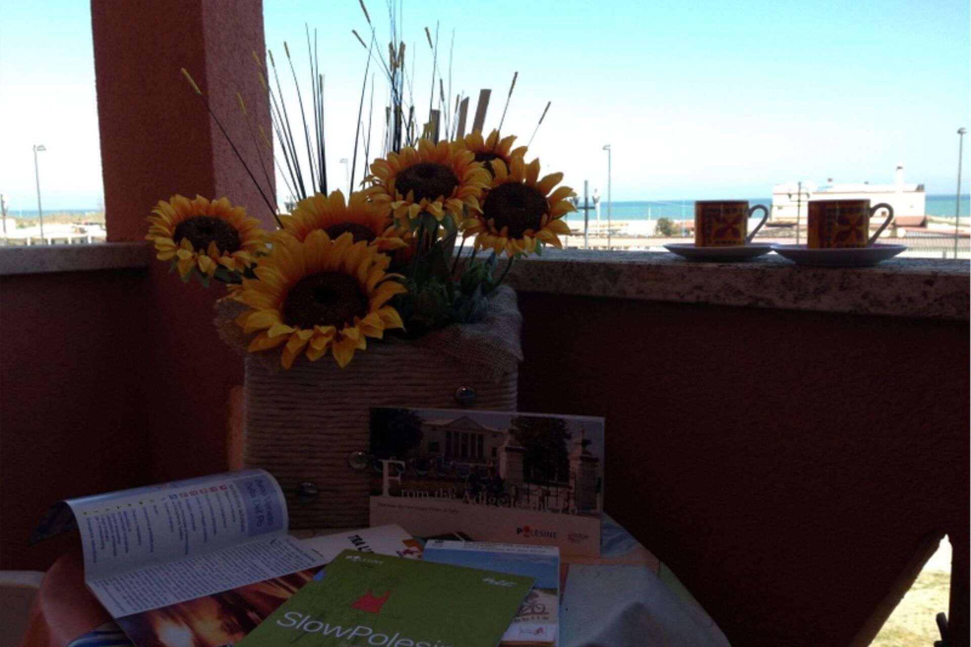 A table with a basket of sunflowers, a book titled 'SlowPoles', and a cup on a balcony overlooking the sea.