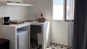 A compact kitchen with a sink, a coffee machine, and a small refrigerator. There is a basket with fruits and a bottle of wine on the counter. - High Point Apartments Fira (Santorini)