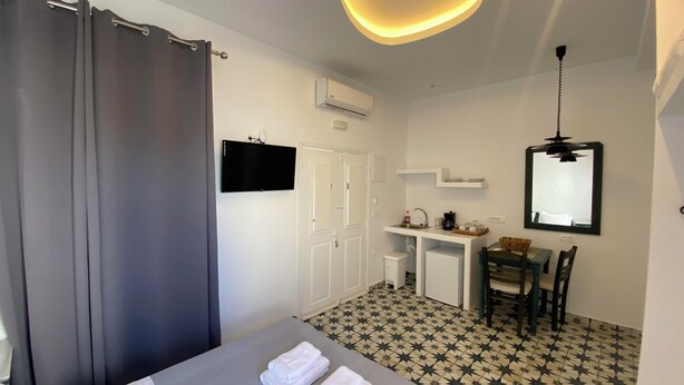 A hotel room with a bed, a desk, a chair, a small table, a mirror, and a ceiling light fixture. - High Point Apartments Fira (Santorini)