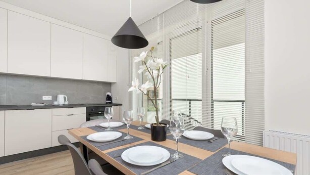 A modern kitchen with white cabinets, a wooden dining table set with white plates and glasses, and a potted orchid on the table.