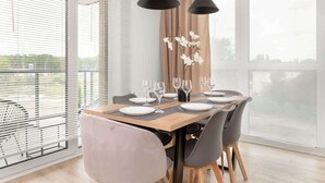 A dining area with a wooden table set for four, surrounded by modern chairs, large windows with blinds, and a view of greenery outside.