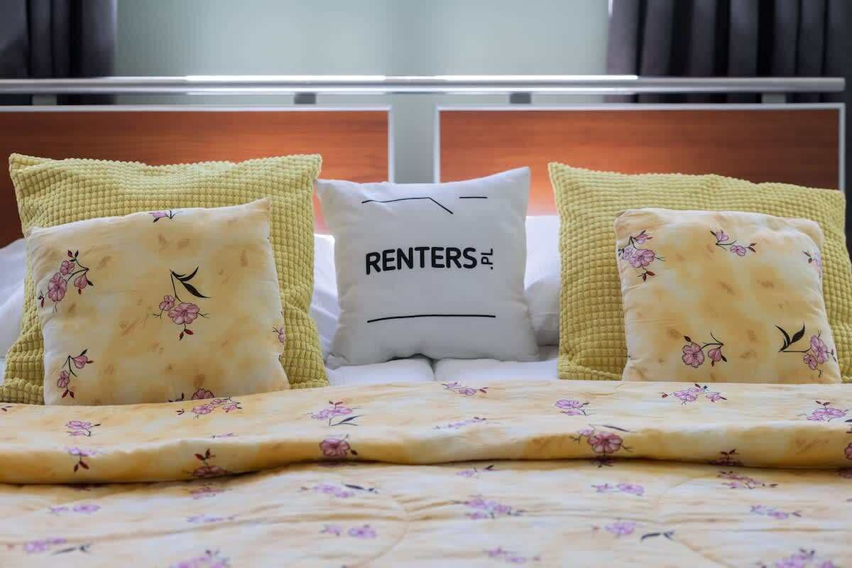 Ein Bett mit floral bedruckter Bettwäsche und Kissen, darunter eines mit der Aufschrift 'RENTERS'.