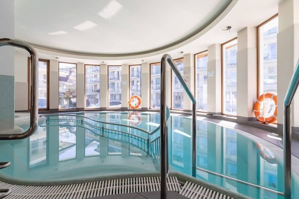 Una piscina interior con paredes de vidrio, barandillas metálicas y ventanas con vistas a la ciudad.