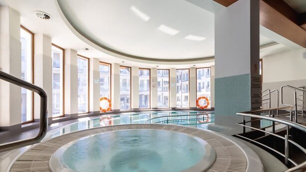 A modern indoor swimming pool with a central hot tub, large windows, and a curved ceiling. - Spacious Apartment | 3 Bedrooms, Pool, Parking. (Zakopane)