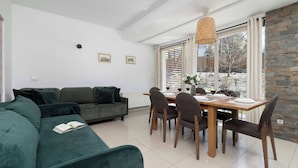 A modern living room with a green sofa, dining table, and chairs, a large window with a view, and a stone wall. - Spacious Apartment | 3 Bedrooms, Pool, Parking. (Zakopane)