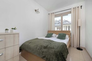 Ein Schlafzimmer mit Bett, Kommode, Lampe und Blick auf Gebäude durch das Fenster.