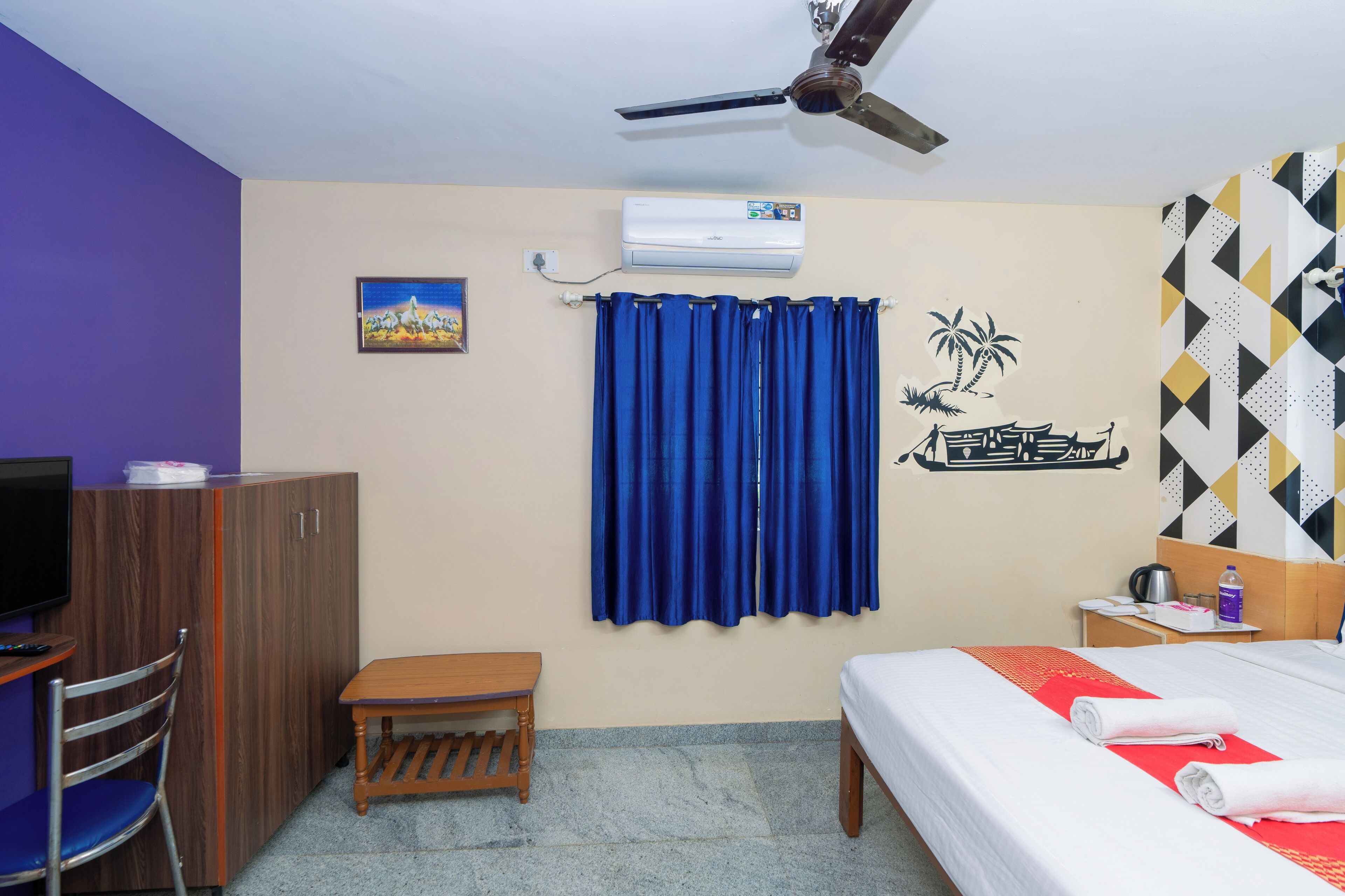 Habitación de hotel con cama, escritorio, silla, ventilador de techo y aire acondicionado, además de un adorno en la pared.