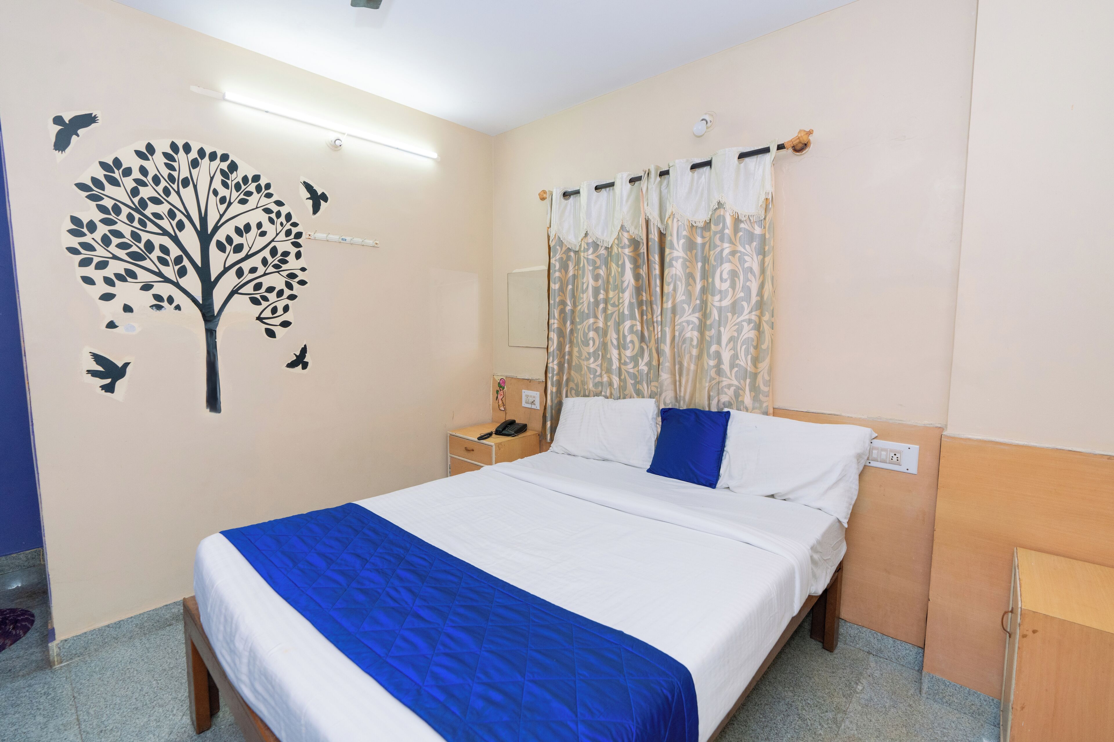 A neatly made bed with a blue bedspread, a wall-mounted tree decal, a nightstand with a phone, and a curtain with a floral pattern.