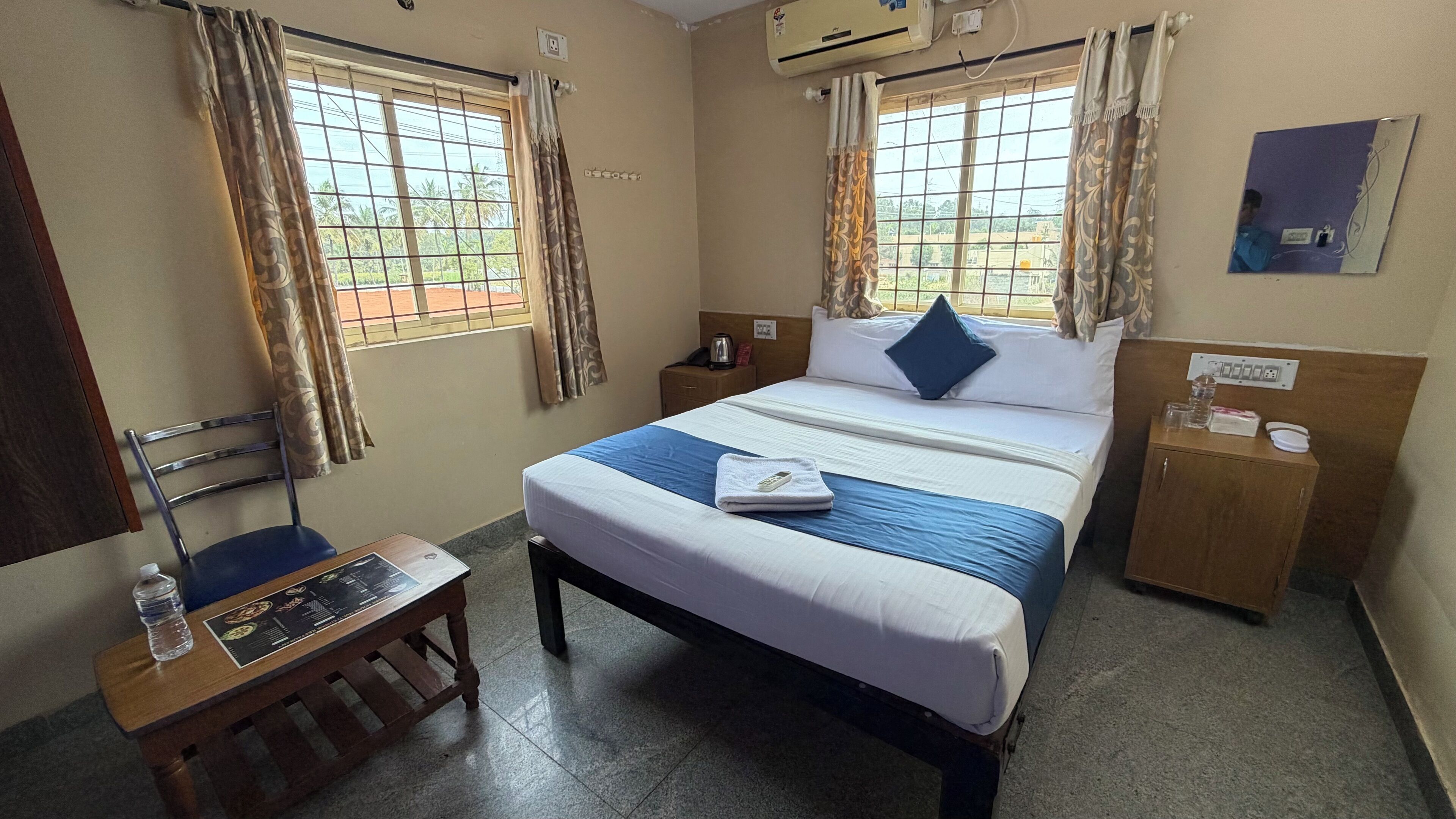A hotel room with a bed, a desk, a chair, a wardrobe, and two windows with curtains.