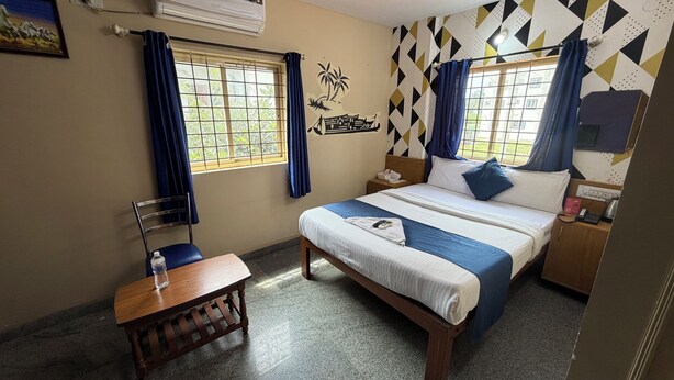 A hotel room with a bed, a desk, a chair, a wall-mounted air conditioner, and a window with curtains. - Hotel Natasha Regency (Bengaluru)