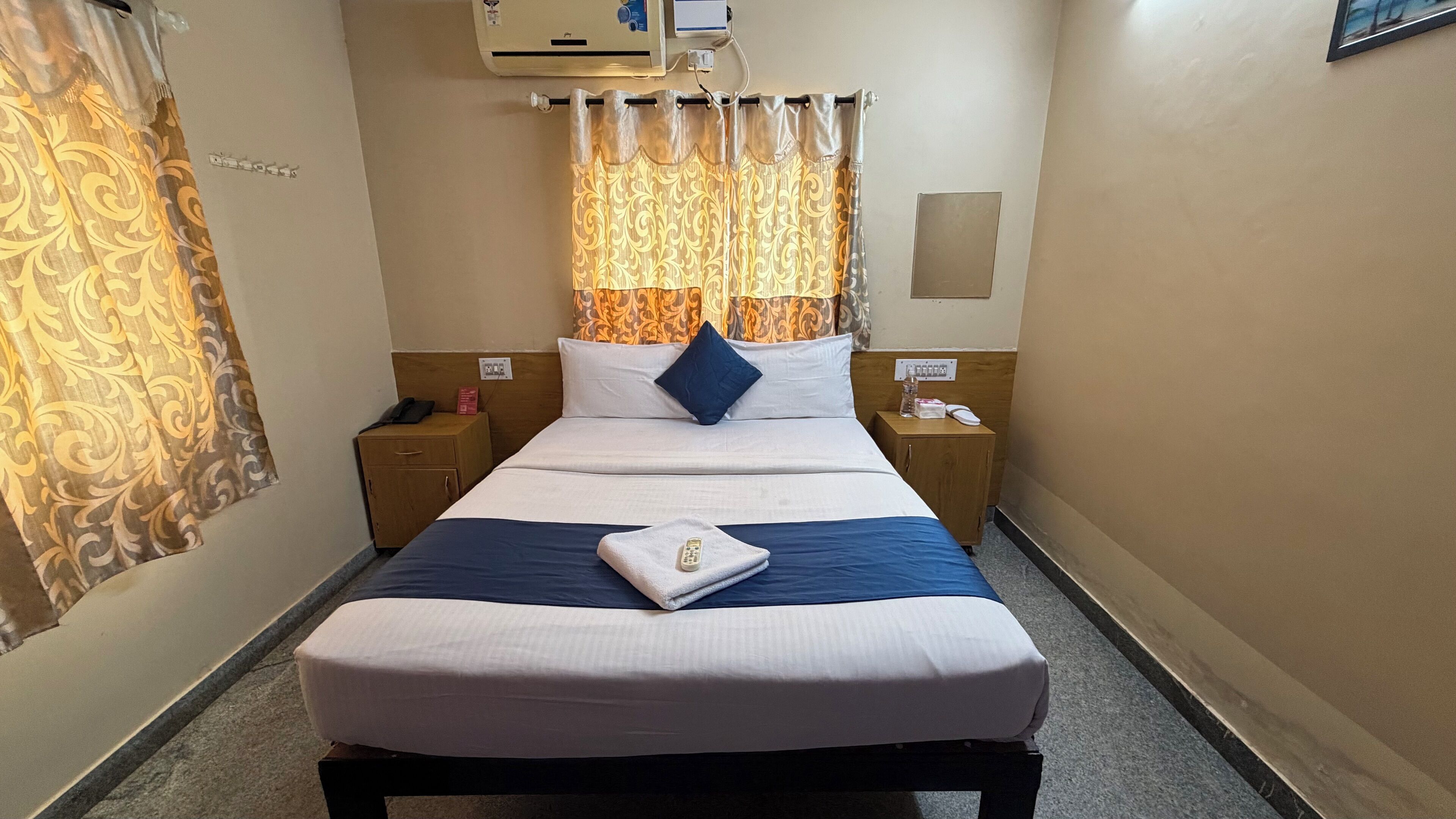 A hotel room with a single bed, a bedside table, a nightstand, a wall-mounted air conditioner, and a curtain with a pattern.