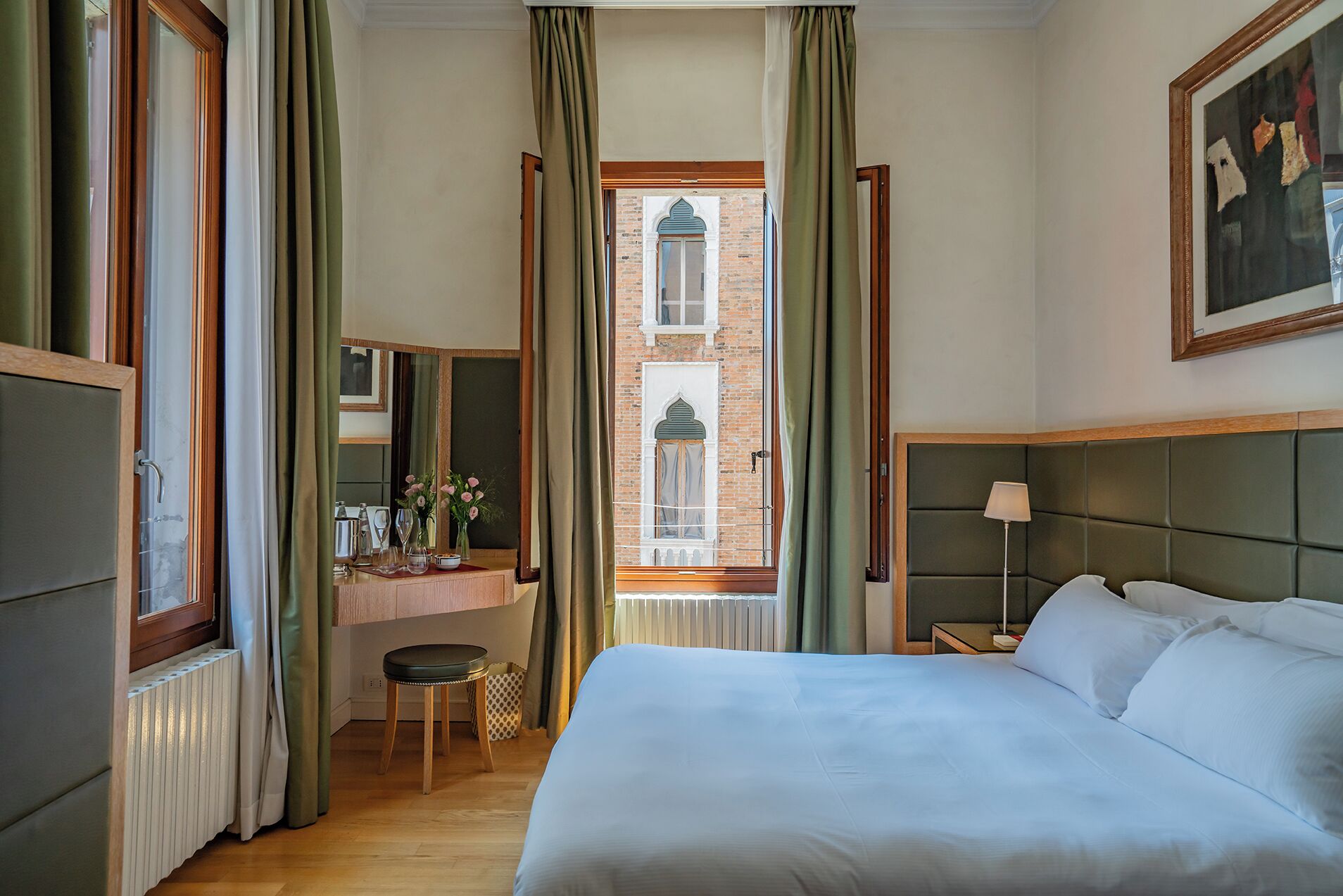 A hotel room with a bed, a desk with a chair, a window with curtains, and a painting on the wall.