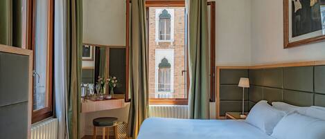 A hotel room with a bed, a desk with a chair, a window with curtains, and a painting on the wall.