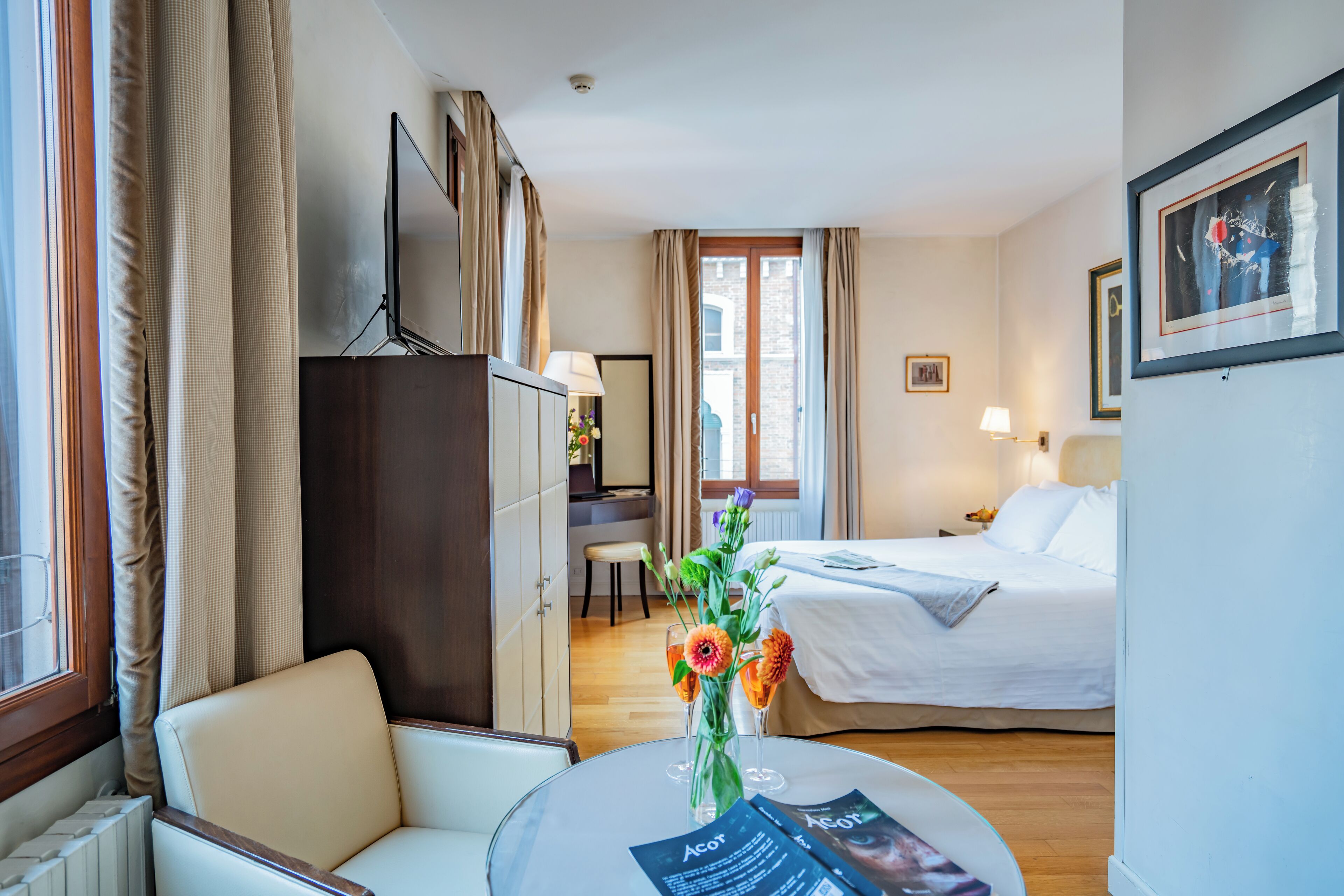 Habitación de hotel con cama, escritorio, silla, una mesita con un florero y un cuadro en la pared.
