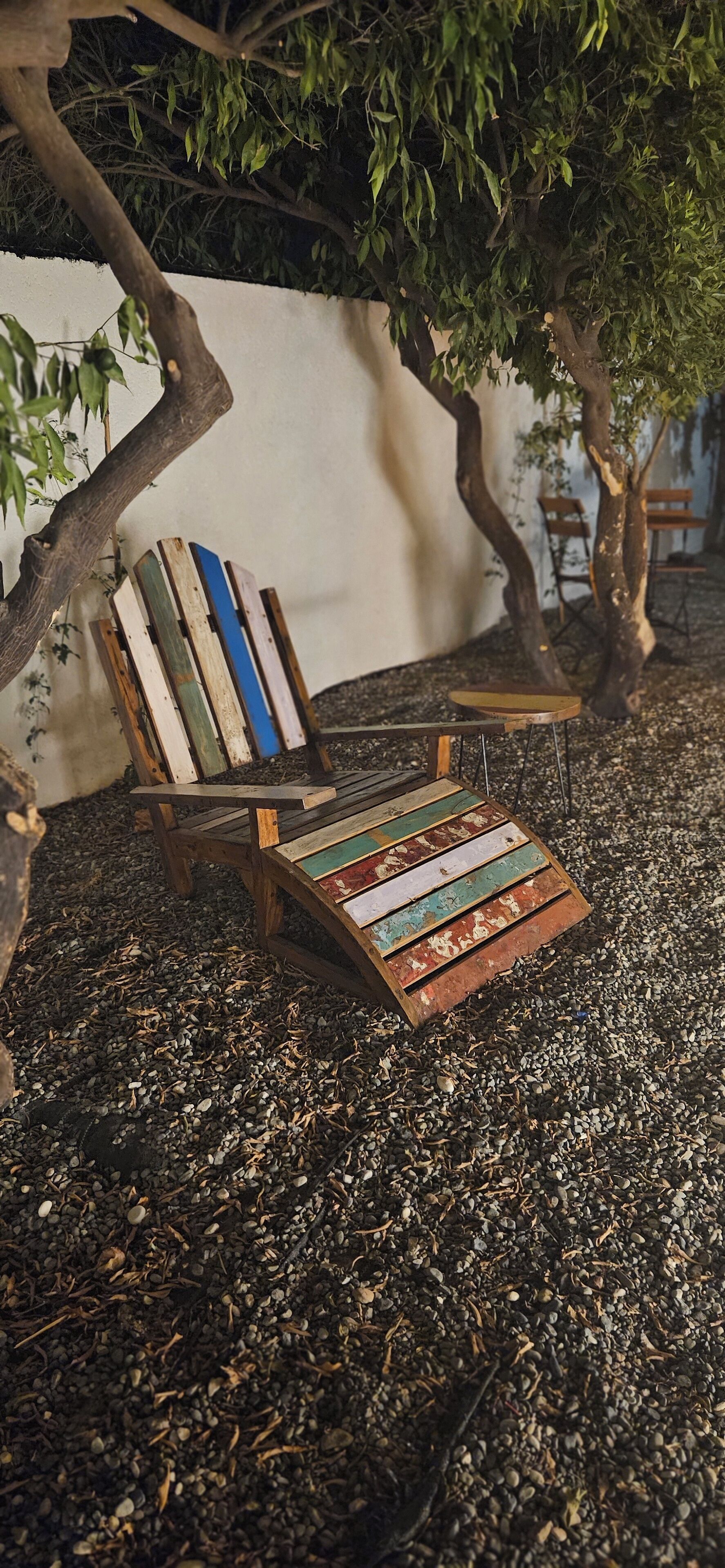 Un banco de madera con un diseño colorido y desgastado, situado sobre grava bajo un árbol.