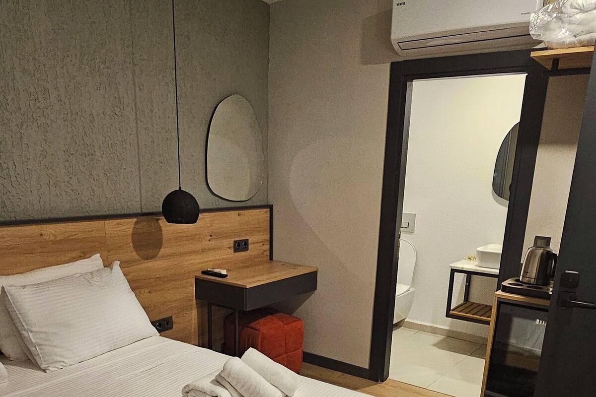 A hotel room with a large bed, a wooden headboard, a bedside table with a remote control, a small table with a kettle, and a mirror on the wall.