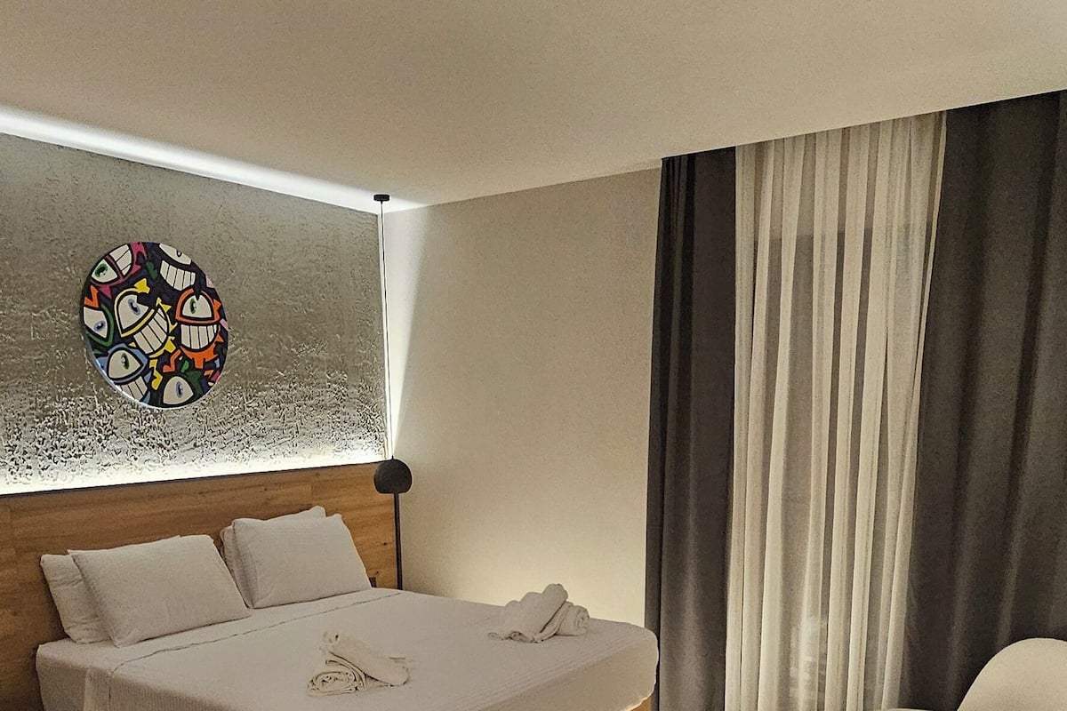 A modern hotel room with a large bed, a bedside table, a lamp, and a view of the city through the window.