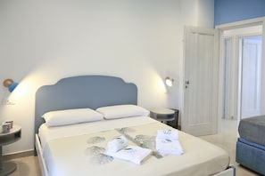Un lit bien fait, avec du linge de lit et des serviettes blanches, une table de chevet avec une lampe, et une porte visible au fond.
