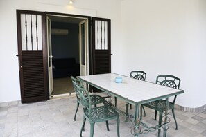 Un coin repas avec une table blanche et quatre chaises vertes, un bol bleu et une porte avec des volets marron.