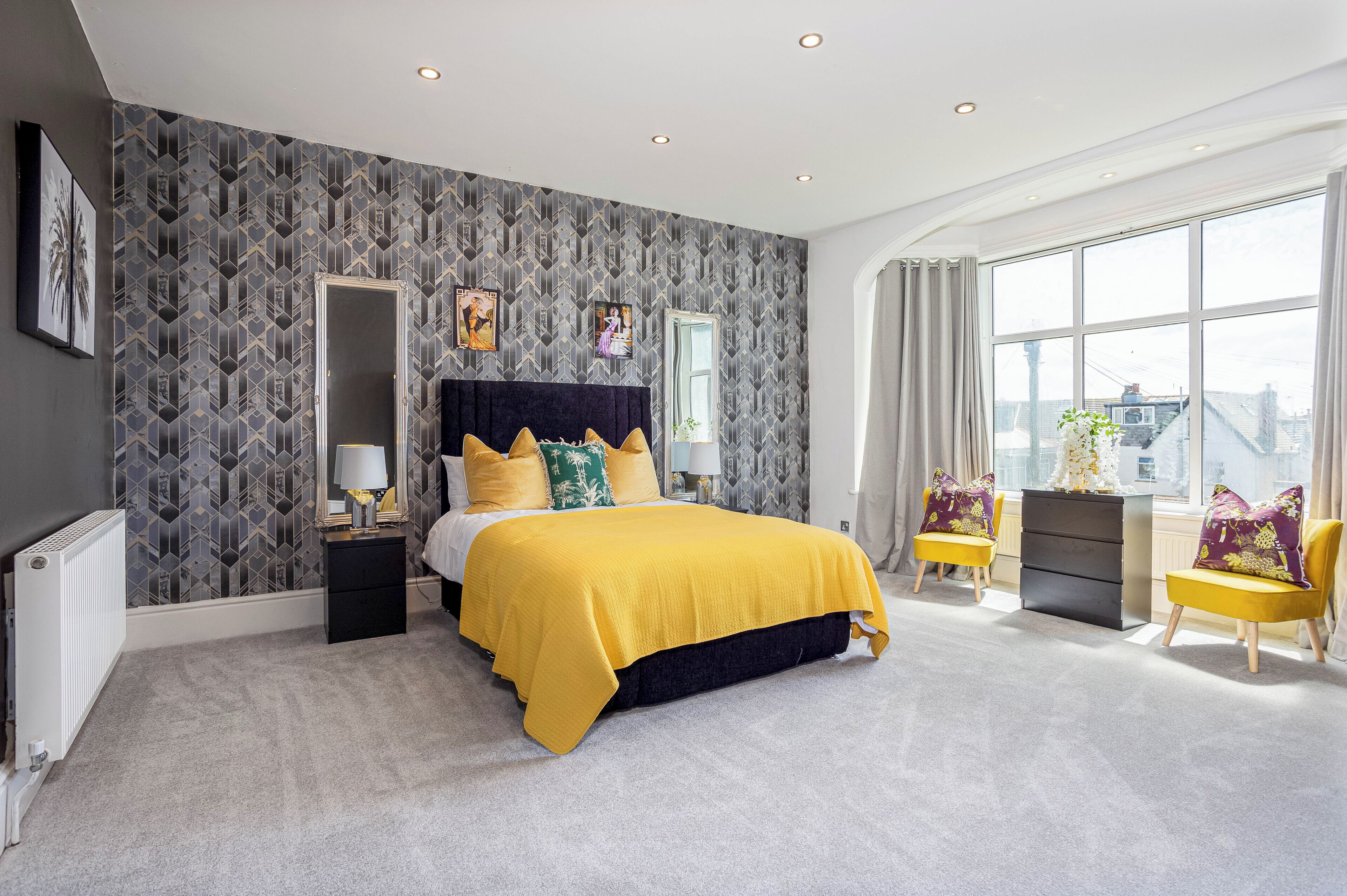 Une chambre à coucher moderne avec un papier peint à motifs géométriques, un grand lit recouvert d’une courtepointe jaune, une table de chevet, une commode et une fenêtre avec des rideaux.