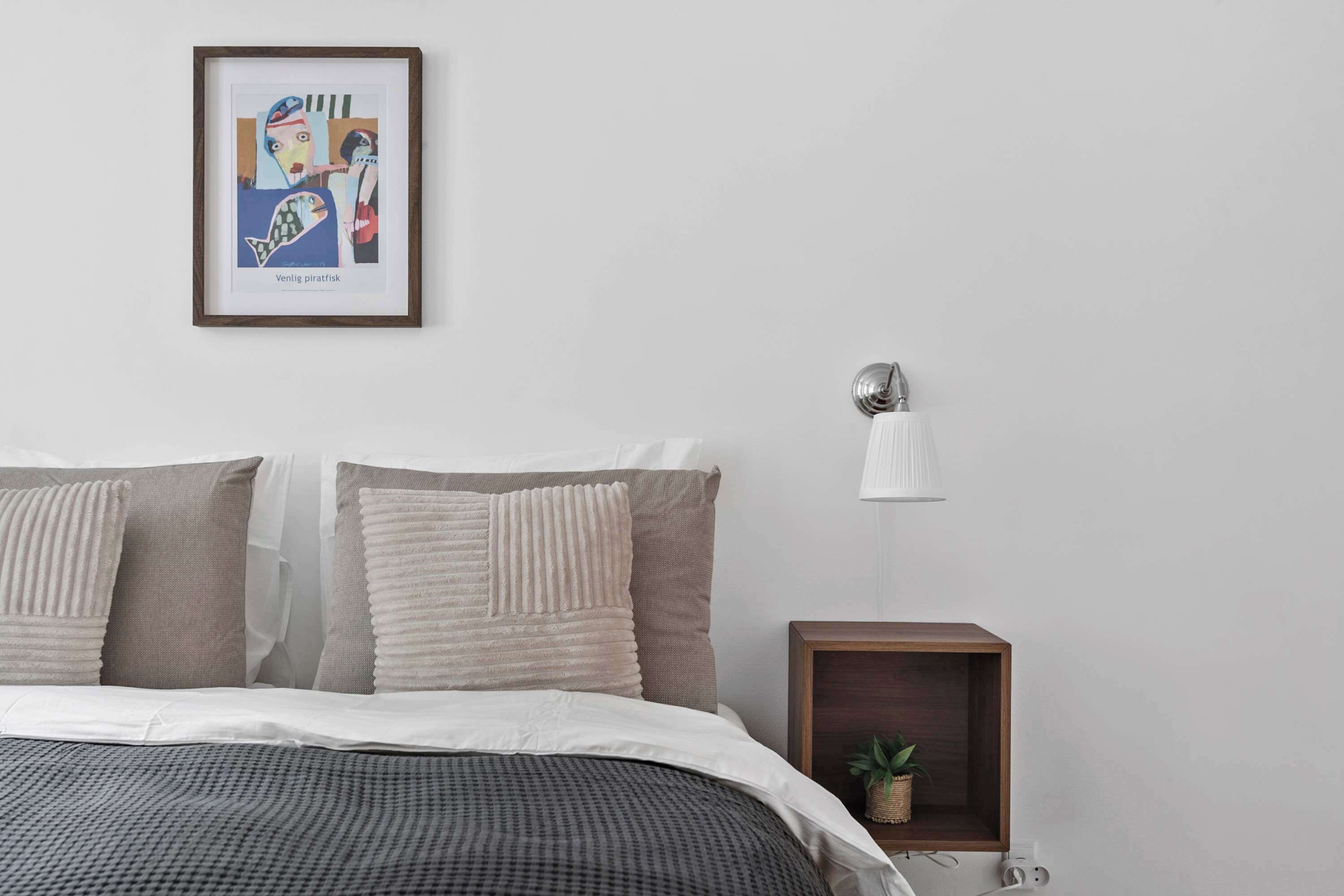 Een slaapkamer met een bed, kussens, een ingelijste afbeelding, een nachtkastje met een lamp en een kleine plant.