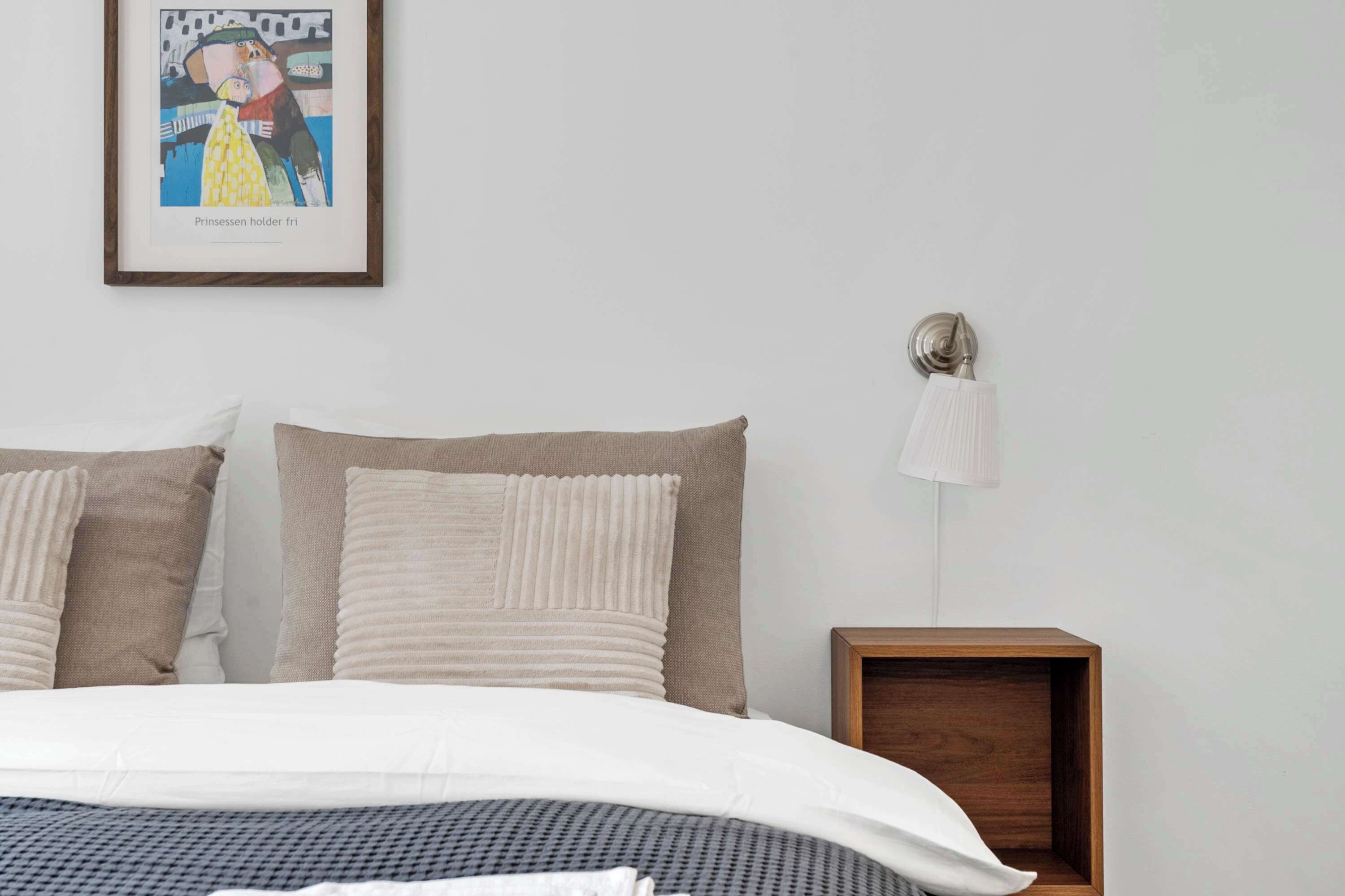 A bedroom with a bed, pillows, a framed picture, a bedside lamp, and a wooden nightstand.