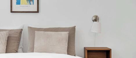 A bedroom with a bed, pillows, a framed picture, a bedside lamp, and a wooden nightstand.