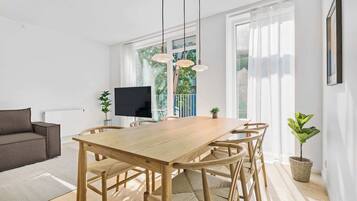 Una sala de estar moderna con una mesa de comedor de madera, sillas de mimbre, un televisor de pantalla plana y ventanas amplias con cortinas translúcidas.