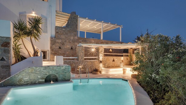 A pool area with a stone wall, a pergola, and a stone building with a light fixture.