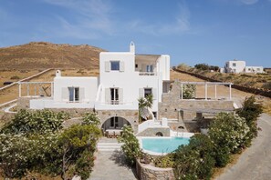 A white villa with a pool, surrounded by greenery and set against a hillside.