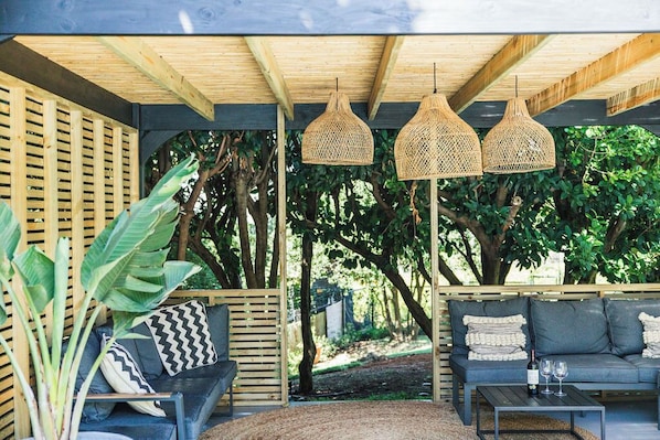 A covered outdoor seating area with wooden beams, woven pendant lights, and a mix of blue and grey sofas.