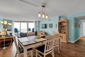 A spacious living area with a dining table, chairs, a sofa, and a TV. There is a bookshelf and a balcony with chairs. - Point unit with Spectacular Views (North Topsail Beach)
