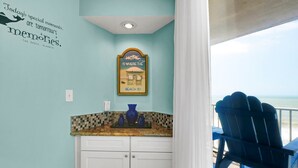 Une salle de bain avec un mur de couleur turquoise, une citation encadrée, un lavabo avec un plan de travail en mosaïque et une chaise bleue près d’une fenêtre donnant sur l’océan.