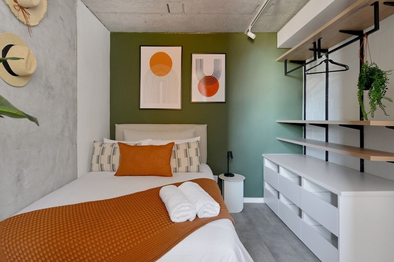 Een moderne slaapkamer met een bed, een nachtkastje, een hangende plant en twee kunstwerken in lijsten aan de muur.
