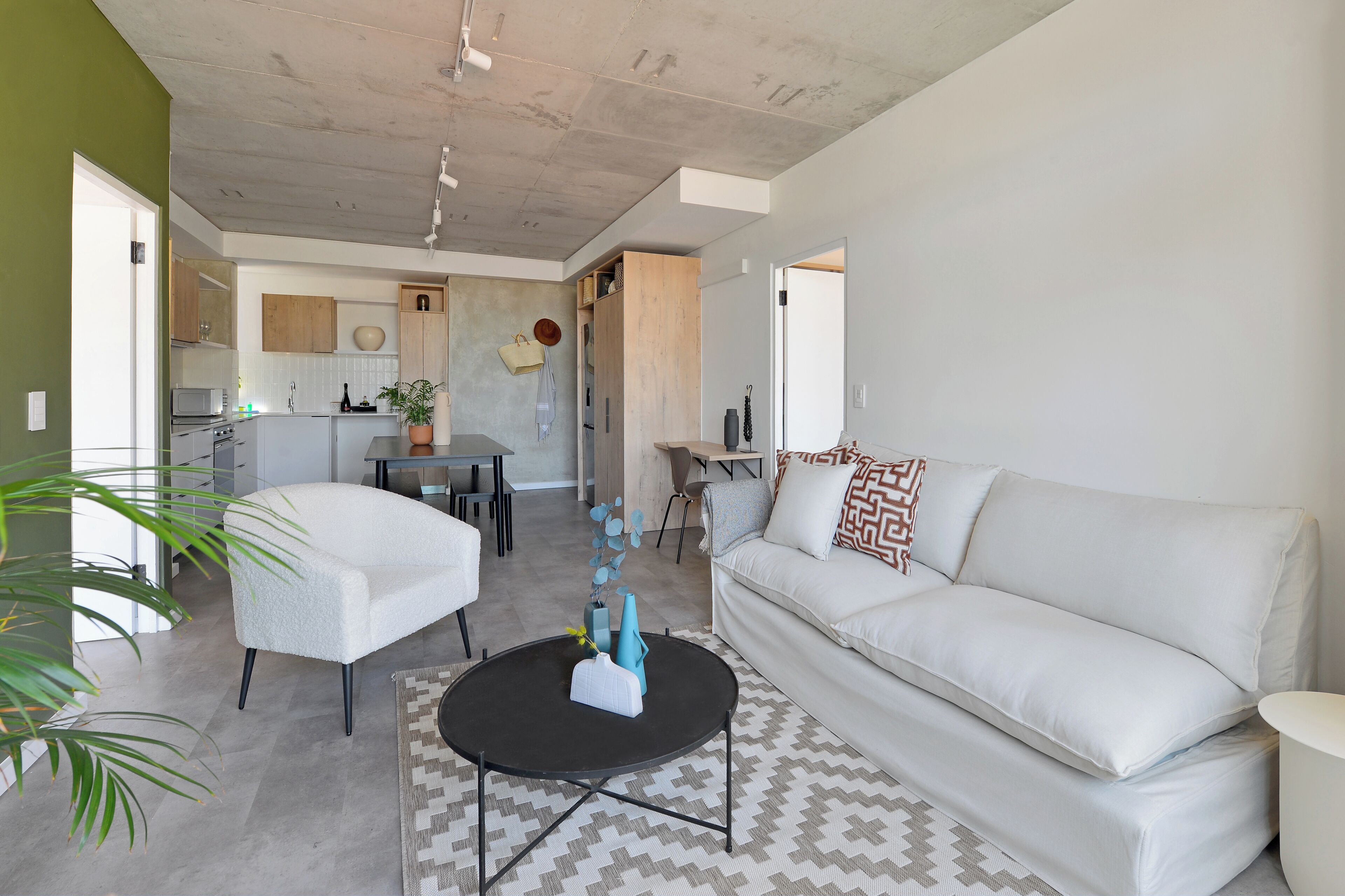 Una sala moderna con un sofá blanco, una mesa de centro negra y un sillón blanco. Se observa una cocina al fondo con armarios de madera y una zona de comedor.