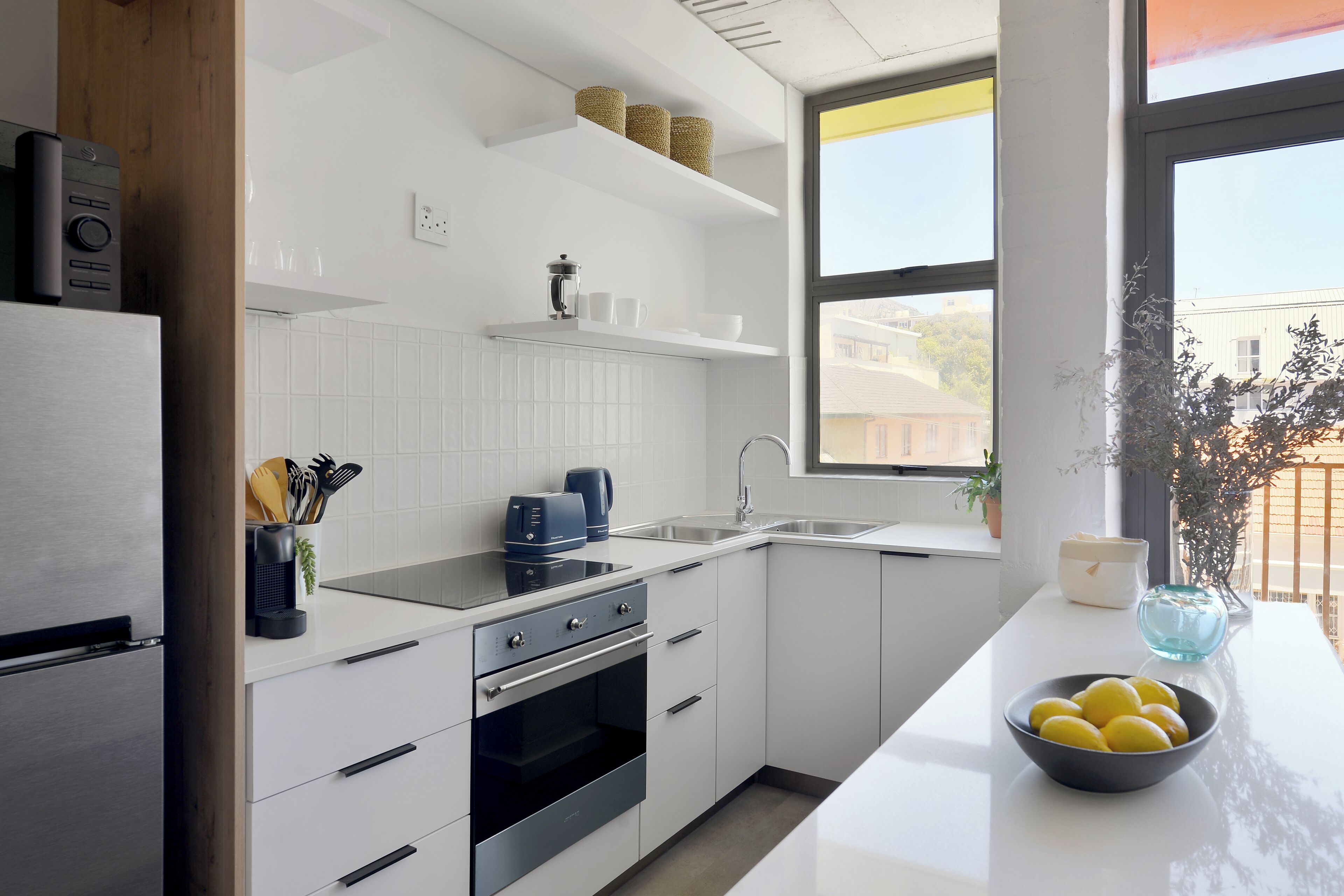 Une cuisine moderne avec des meubles blancs, un réfrigérateur en acier inoxydable, un four encastré et un évier. On trouve des paniers sur l’étagère supérieure et un bol de citrons sur le plan de travail.