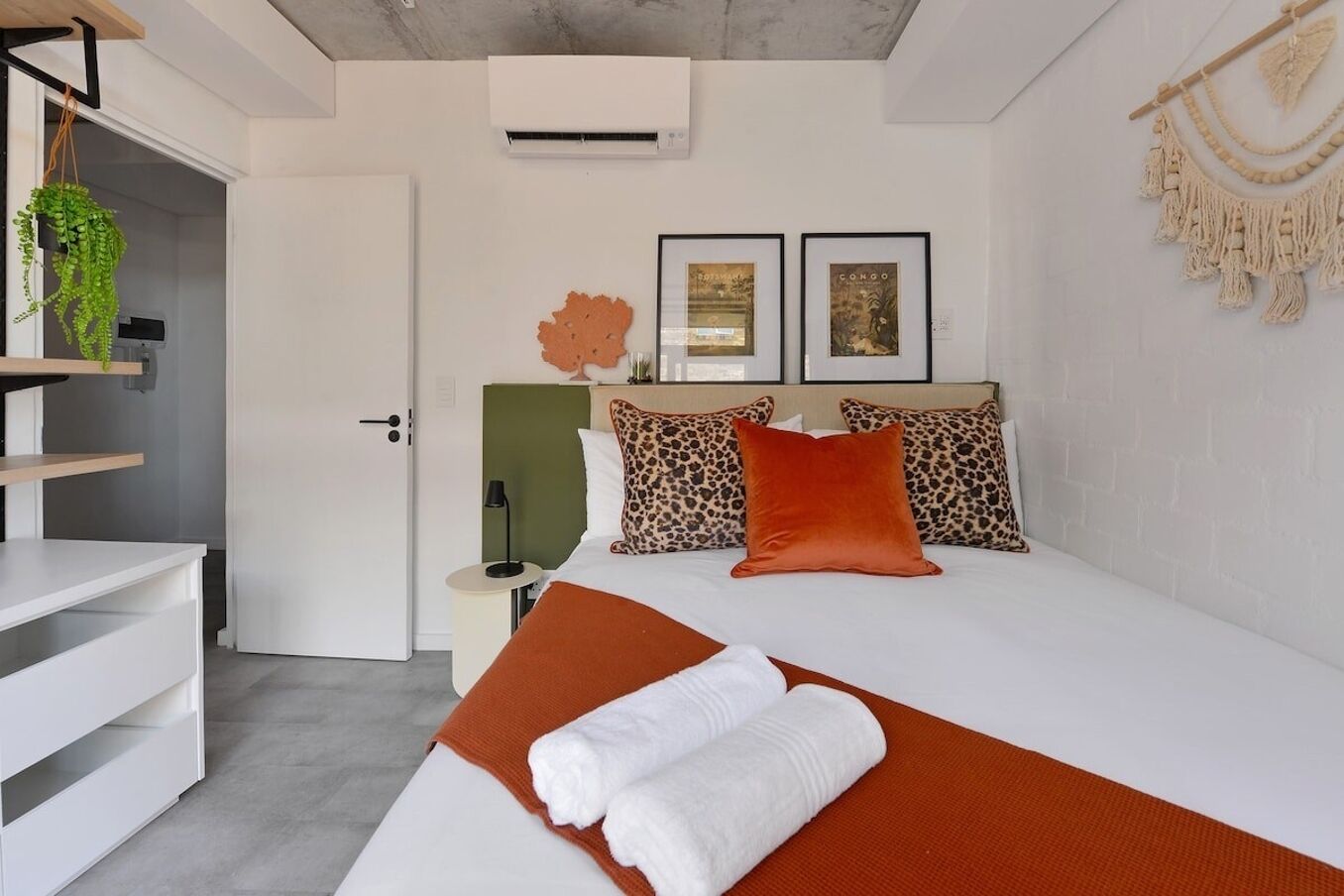 A bedroom with a bed, a nightstand, a lamp, a plant, and two framed pictures on the wall.