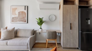 Un salon moderne comprenant un canapé, un petit bureau, une chaise, une plante en pot, une horloge murale et un meuble encastré en bois avec un réfrigérateur.