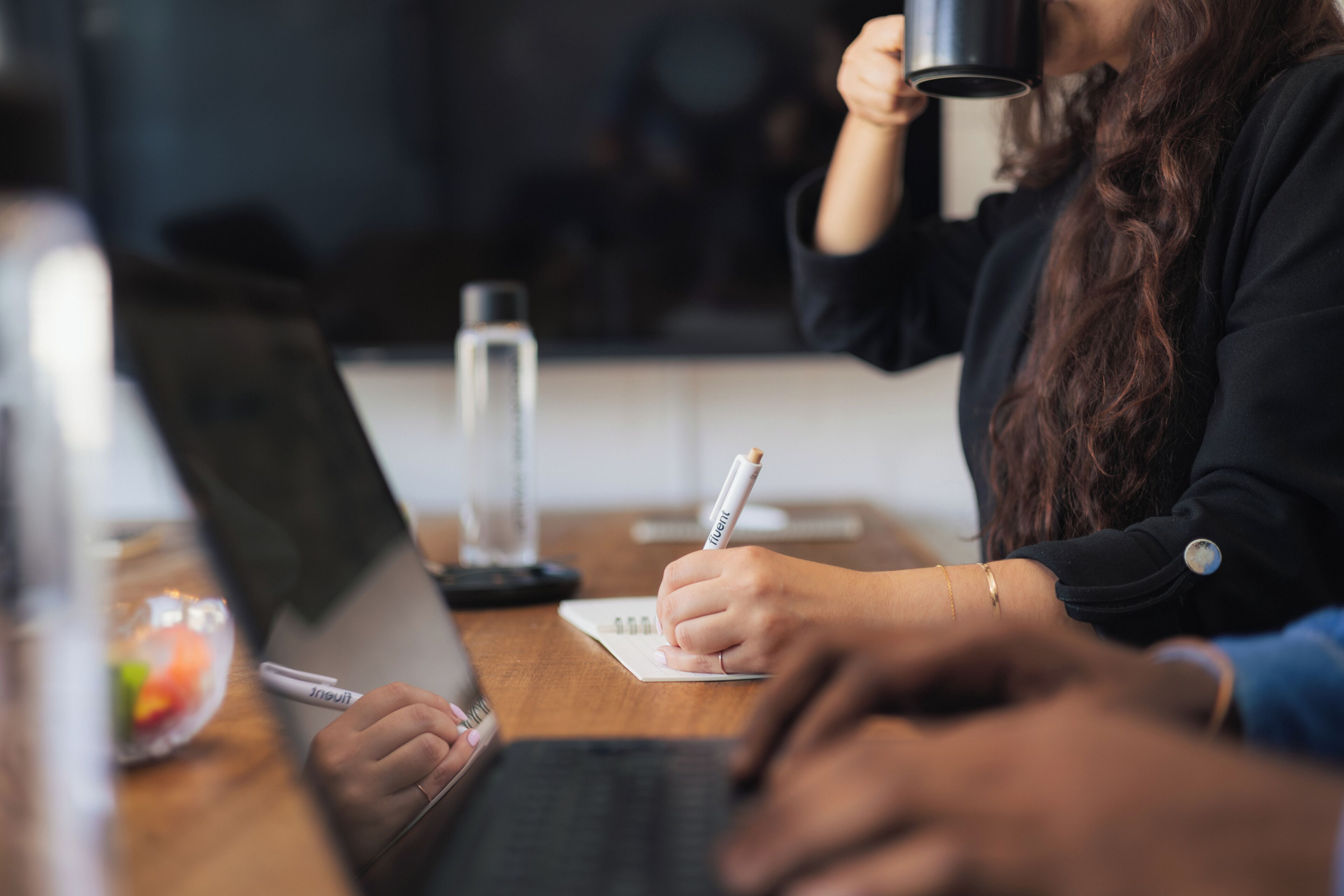 A person writing on a notepad with a pen, a laptop, and a smartphone on a wooden table.