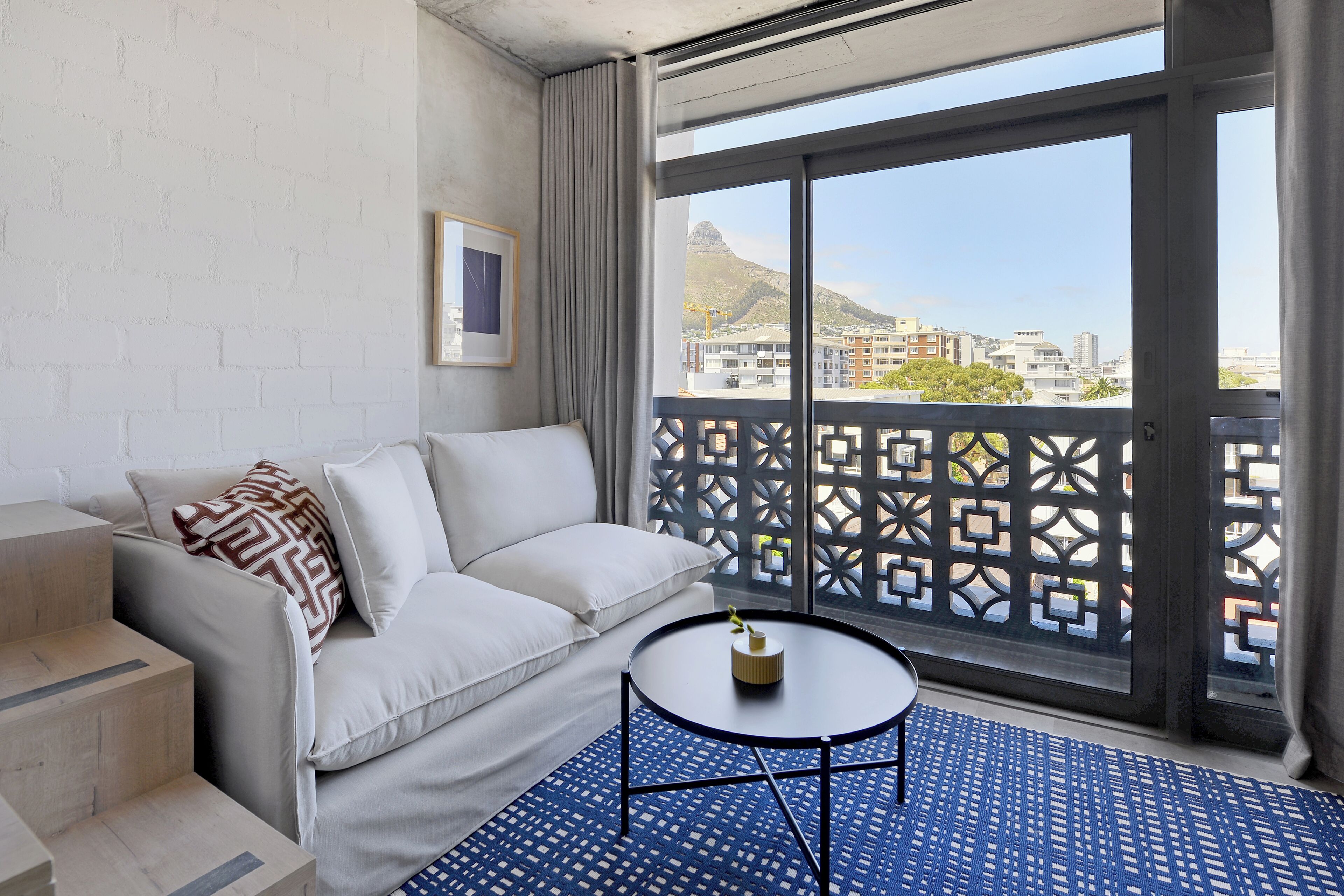 Una habitación de hotel moderna con balcón, un sofá blanco, una mesita redonda pequeña y una alfombra azul estampada.