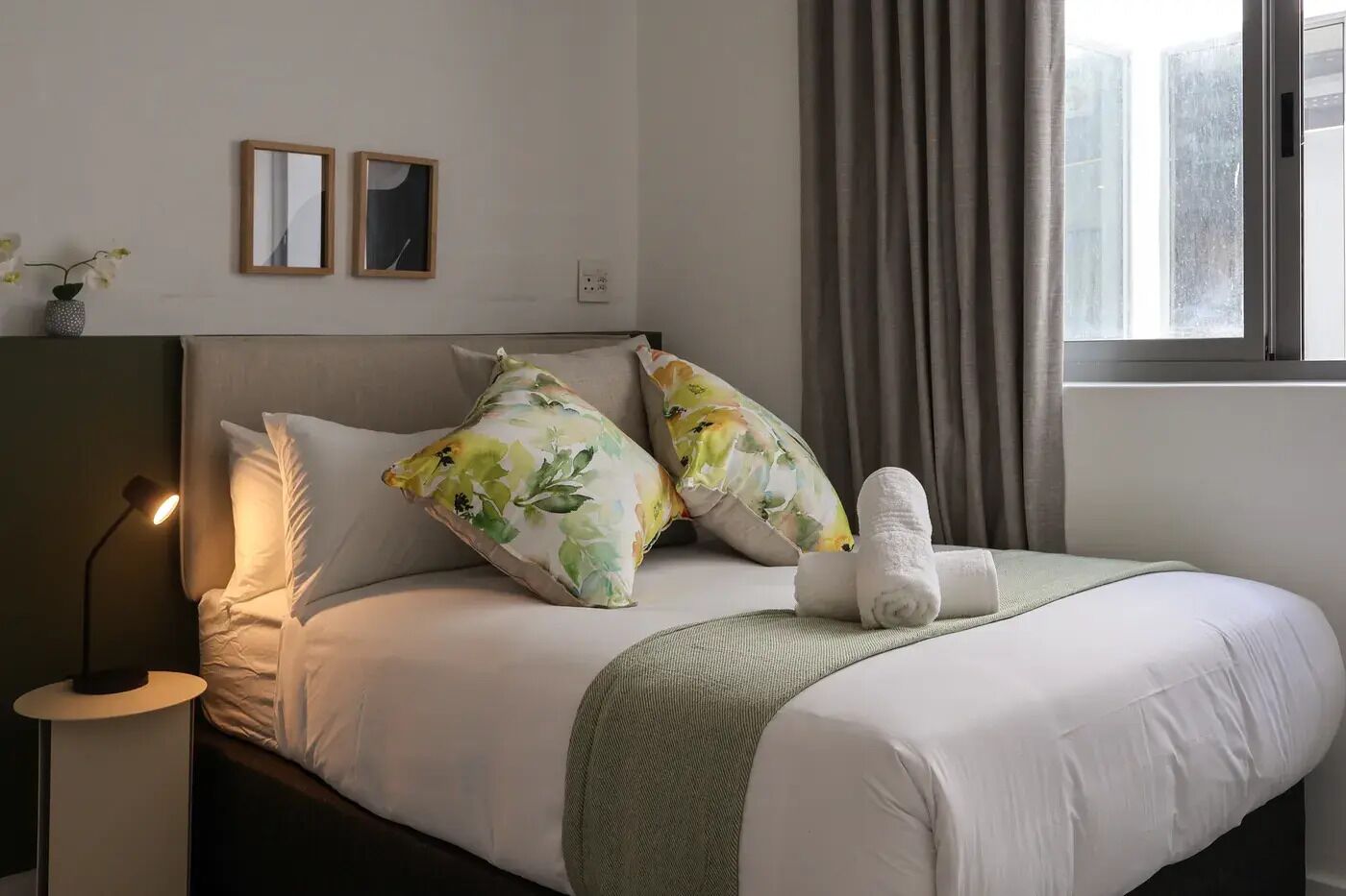 A hotel room with a bed, pillows, a bedside lamp, a window with curtains, and a wall-mounted picture.